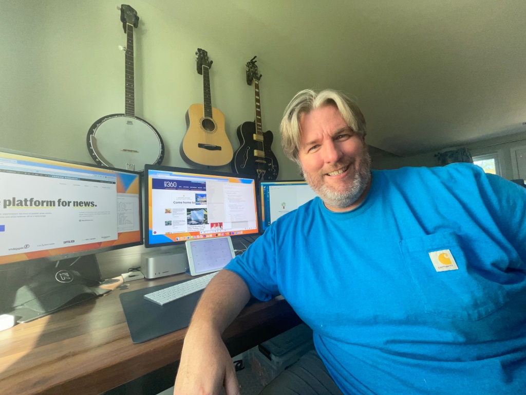 Daniel Brown at his home-based workstation. Dan is relaxed, leaning against his desk, smiling broadly and wearing a tee shirt. Three monitors, a tablet, and an Apple Studio are visible. Behind him, on the wall, hang buitars and a banjo.